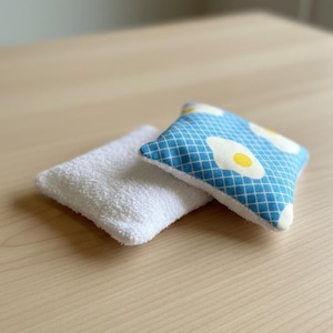 May include: Two small, square fabric items rest on a light wood surface. One is white and fluffy, the other is blue with a white grid pattern and egg graphics. The egg-themed item has a yellow yolk.