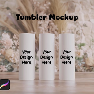May include: Three white tumblers with black text that reads "Your Design Here" on a wooden surface with a blurred background of beige flowers.