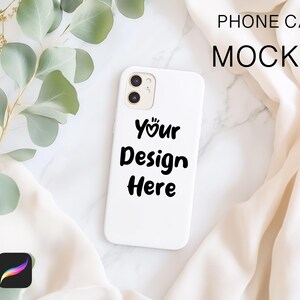 May include: White phone case mockup with the text "Your Design Here" on a white marble surface with a white fabric and green leaves.