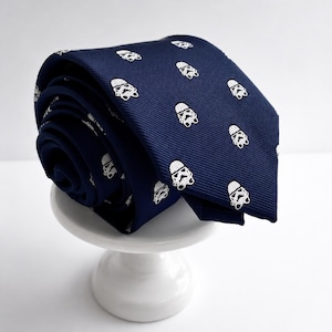 May include: A navy blue necktie featuring a repeating white pattern of Stormtrooper helmets. The fabric has a subtle ribbed texture, and the tie is rolled into a circular shape, displaying the pointed end and the consistent graphic print throughout.
