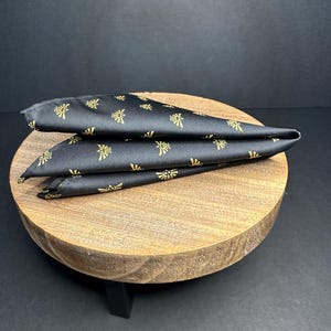 May include: A black pocket square with a gold pattern of a stylized triangle with a bird inside. The pocket square is folded and sitting on a wooden stand.