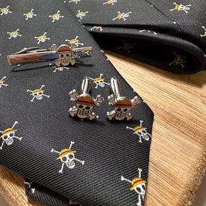 May include: A black tie, tie clip, and cufflinks set with a repeating pattern of white skulls with crossbones and yellow hats. The tie clip and cufflinks have a skull design with a red stripe.