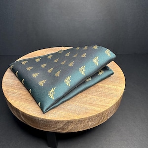 Op de afbeelding: Een donkergroene zakdoek met een gouden patroon van een gestileerde vogel. De zakdoek is gevouwen en wordt getoond op een houten standaard.