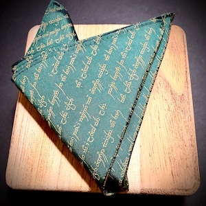 Könnte beinhalten: Ein gefaltetes grünes Einstecktuch mit goldener Schrift, das auf einer Holzoberfläche liegt. Der Stoff hat ein sich wiederholendes Muster aus stilisiertem Text. Die Kanten sind sauber mit goldenem Faden genäht, was ein sauberes, maßgeschneidertes Aussehen ergibt.
