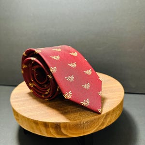 Könnte beinhalten: Eine aufgerollte bordeauxfarbene Krawatte mit einem sich wiederholenden goldenen Emblem-Muster. Die Krawatte wird auf einem kleinen, runden Holzständer präsentiert. Der Hintergrund ist dunkel. Ein klassisches Accessoire.