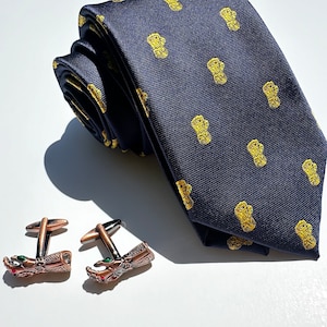 May include: A dark blue silk tie with a repeating pattern of small gold figures. Two cufflinks with a detailed design of a hand with red and green gemstones.