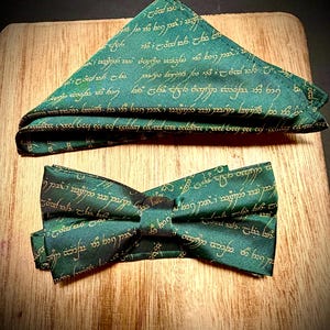 Puede incluir: Un lazo y pañuelo de bolsillo verdes con texto dorado. La tela tiene un patrón repetido de escritura. El lazo está atado y el pañuelo está doblado en un triángulo. Sobre una superficie de madera.