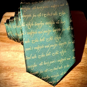 Può includere: Una cravatta verde con scritte dorate, probabilmente a tema fantasy. La cravatta è piegata e appoggiata su una superficie di legno. Le scritte dorate formano un motivo ripetuto.