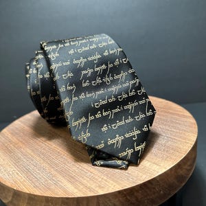 May include: Black necktie with gold-colored script. The tie is folded and rests on a small wooden stand. The script appears to be a stylized font. The tie is a dark color, and the script is a contrasting gold.