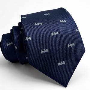 May include: A navy blue necktie featuring a repeating pattern of small, light gray bat-shaped icons. The fabric has a diagonal ribbed texture, and the tie is folded in a loose coil, displaying the pointed tip and the main body of the accessory.