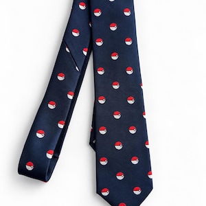 May include: A navy blue necktie featuring a repeating pattern of small, circular red and white Poké Ball icons. The tie has a pointed tip and a smooth, woven texture, displaying a consistent print of the spherical gaming-themed symbols throughout.