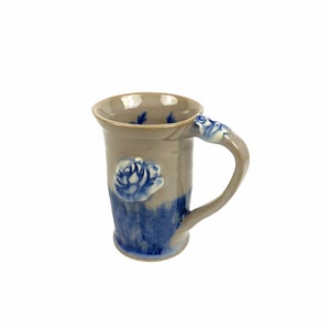Può includere: Una tazza in ceramica grigia con un motivo floreale blu e bianco. La tazza ha un manico e una base arrotondata.