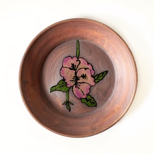 Può includere: Un piatto in ceramica marrone con un disegno di fiore rosa dipinto a mano. Il fiore ha foglie verdi e un contorno nero.