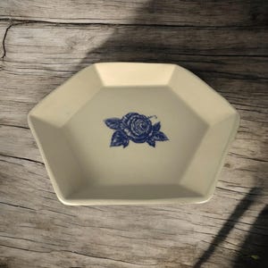 Op de afbeelding: Een wit zeshoekig keramisch schaaltje met een blauw bloemmotief van een roos. Het schaaltje is perfect om kleine snacks te serveren of om sieraden in te bewaren.