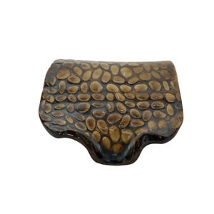 Può includere: Un piatto in ceramica marrone con una superficie testurizzata che ricorda piccoli ciottoli. Il piatto ha una forma irregolare con un bordo leggermente ondulato.