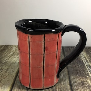 May include: A handmade ceramic mug with a black rim and handle. The mug is decorated with a red and black checkered pattern.