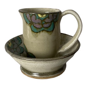 Può includere: Un set di tazza e piattino in ceramica fatto a mano con un design floreale in tonalità di verde, viola e giallo. La tazza ha un manico e il piattino è rotondo.