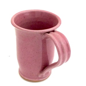 May include: A pink ceramic mug with a wide, rounded base and a curved handle. The mug has a speckled glaze.