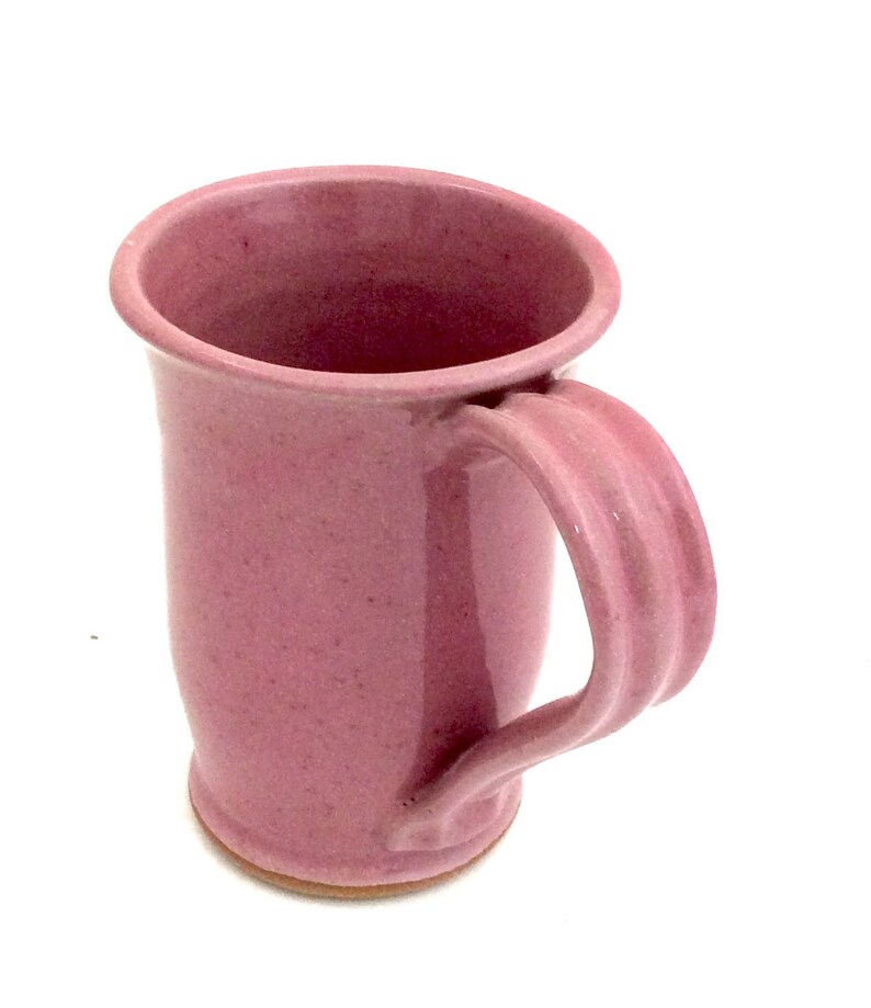 May include: A pink ceramic mug with a wide, rounded base and a curved handle. The mug has a speckled glaze.