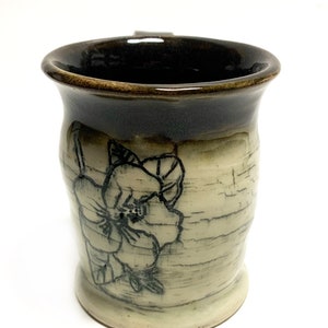 Può includere: Una tazza in ceramica con una glassa marrone e un design floreale bianco. La tazza ha una forma arrotondata e un manico.