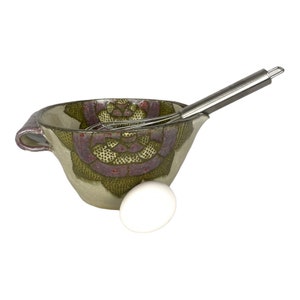 Può includere: Una ciotola per mescolare in ceramica con un motivo floreale in tonalità di verde, viola e rosso. Una frusta d'argento è appoggiata nella ciotola e un uovo bianco è posto davanti alla ciotola.