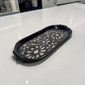May include: A black ceramic tray with a floral design. The tray is oblong with rounded ends and a slightly raised rim. The interior features a pattern of white daisy-like flowers against a dark background. The tray is suitable for serving or display.