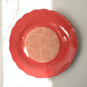 Può includere: Un piatto in ceramica color corallo con un bordo ondulato. Il centro del piatto presenta un cerchio bianco con un delicato motivo floreale.