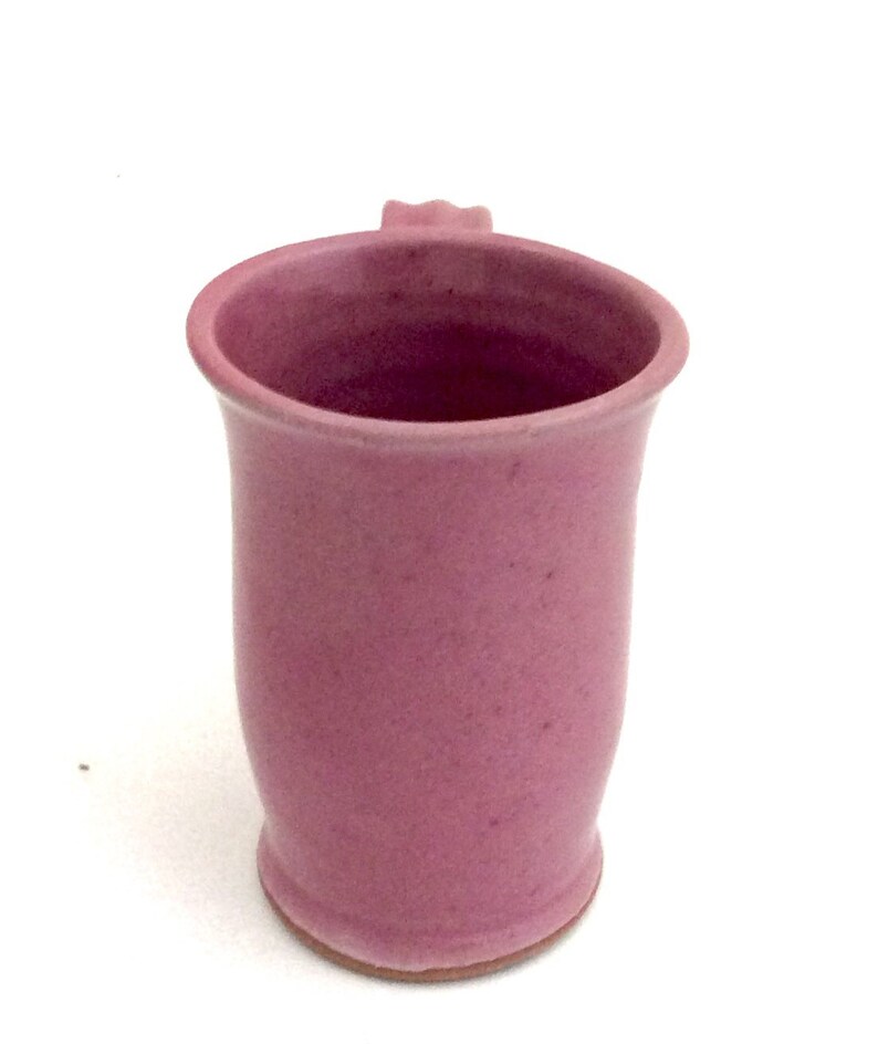 May include: A pink ceramic mug with a rounded shape and a small handle. The mug has a smooth, slightly textured surface.