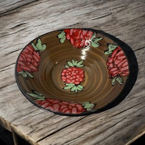 Può includere: Una ciotola in ceramica marrone con un bordo nero, decorata con motivi floreali rossi e verdi. La ciotola ha un motivo a spirale all'interno.
