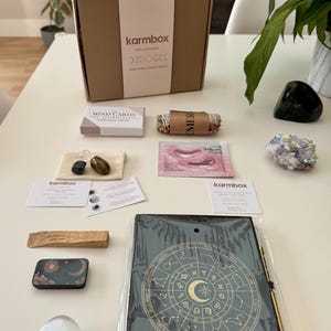 May include: A Karmbox subscription box set containing a cardboard box, a deck of Mind Cards, a sage bundle, a small pouch with three crystals, a Japan Sakura face mask, two informational cards, a palo santo stick, a metal tin, a crystal, and a zodiac journal.