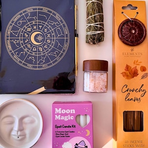 May include: A collection of items including a dark blue zodiac journal, a bundle of dried herbs, a small glass jar with pink salt, a white ceramic face candle holder, a pink Moon Magic spell candle kit box, and an orange Crunchy Leaves incense box.
