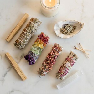 May include: An assortment of sage bundles, palo santo sticks, and a lit candle. The bundles are adorned with colorful flowers and berries. A seashell with herbs, matches, and a selenite wand complete the arrangement.