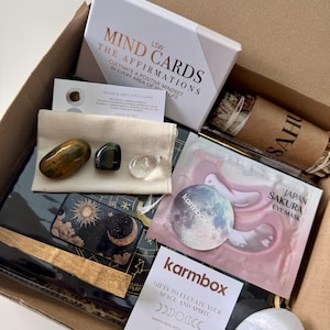 May include: A cardboard gift box containing LSW Mind Cards, a Japan Sakura eye mask, a bundle of sage, a white card with crystal information, a small cream pouch with three polished stones, a black celestial-themed journal, and a white stone.