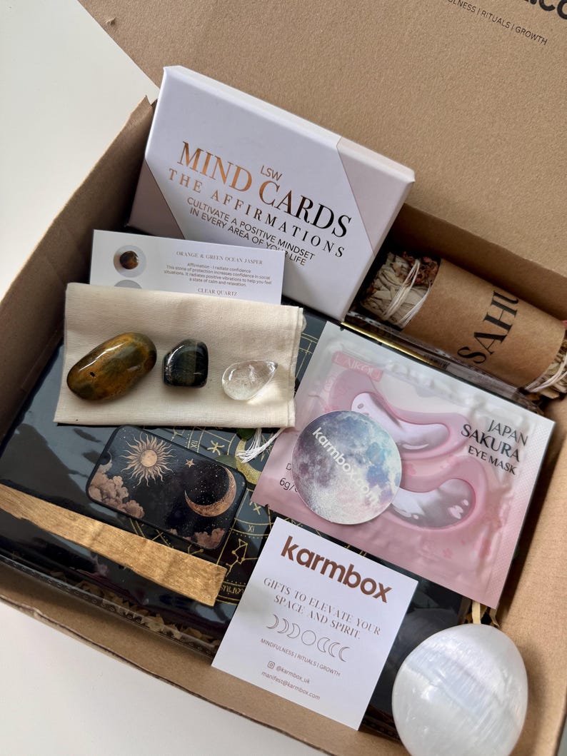 May include: A cardboard gift box containing LSW Mind Cards, a Japan Sakura eye mask, a bundle of sage, a white card with crystal information, a small cream pouch with three polished stones, a black celestial-themed journal, and a white stone.