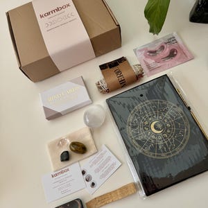 May include: A brown cardboard Karmbox subscription box with a light pink sleeve, a deck of Mind Cards, a clear crystal sphere, two small polished stones in a beige pouch, a black zodiac journal, a sage bundle, a Japan Sakura eye mask, and a small tin.