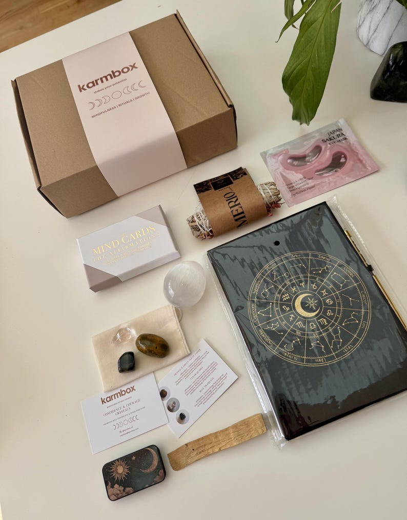 May include: A brown cardboard Karmbox subscription box with a light pink sleeve, a deck of Mind Cards, a clear crystal sphere, two small polished stones in a beige pouch, a black zodiac journal, a sage bundle, a Japan Sakura eye mask, and a small tin.