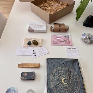 May include: A Karmbox subscription box containing a deck of Mind Cards, a sage bundle, pink Sakura eye masks, three tumbled crystals in a pouch, two informational cards, a palo santo stick, a celestial tin, a moon sticker, a crystal egg, and a zodiac journal.
