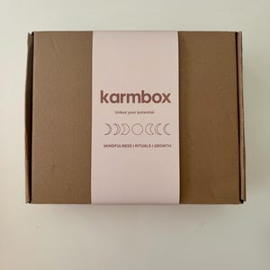 May include: A rectangular brown cardboard box featuring a light pink paper sleeve. The sleeve displays the text "karmbox", "Unbox your potential", and "MINDFULNESS | RITUALS | GROWTH", along with a graphic illustration of moon phases.