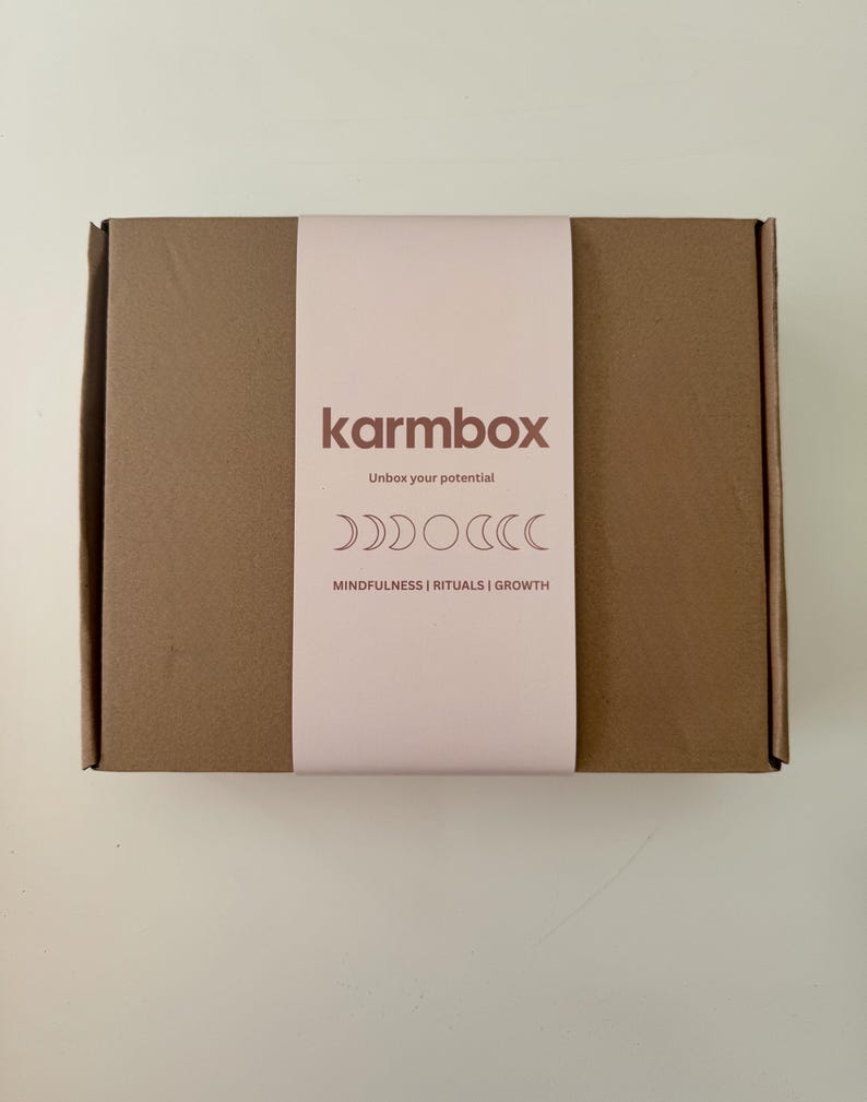 May include: A rectangular brown cardboard box featuring a light pink paper sleeve. The sleeve displays the text "karmbox", "Unbox your potential", and "MINDFULNESS | RITUALS | GROWTH", along with a graphic illustration of moon phases.
