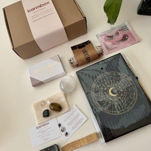 May include: A Karmbox cardboard shipping box, a pack of Japan Sakura eye masks, a sage bundle, a Mind Cards deck, a selenite stone, a small pouch with three crystals, a Karmbox card, a black zodiac journal, a palo santo stick, and a small sun and moon tin.