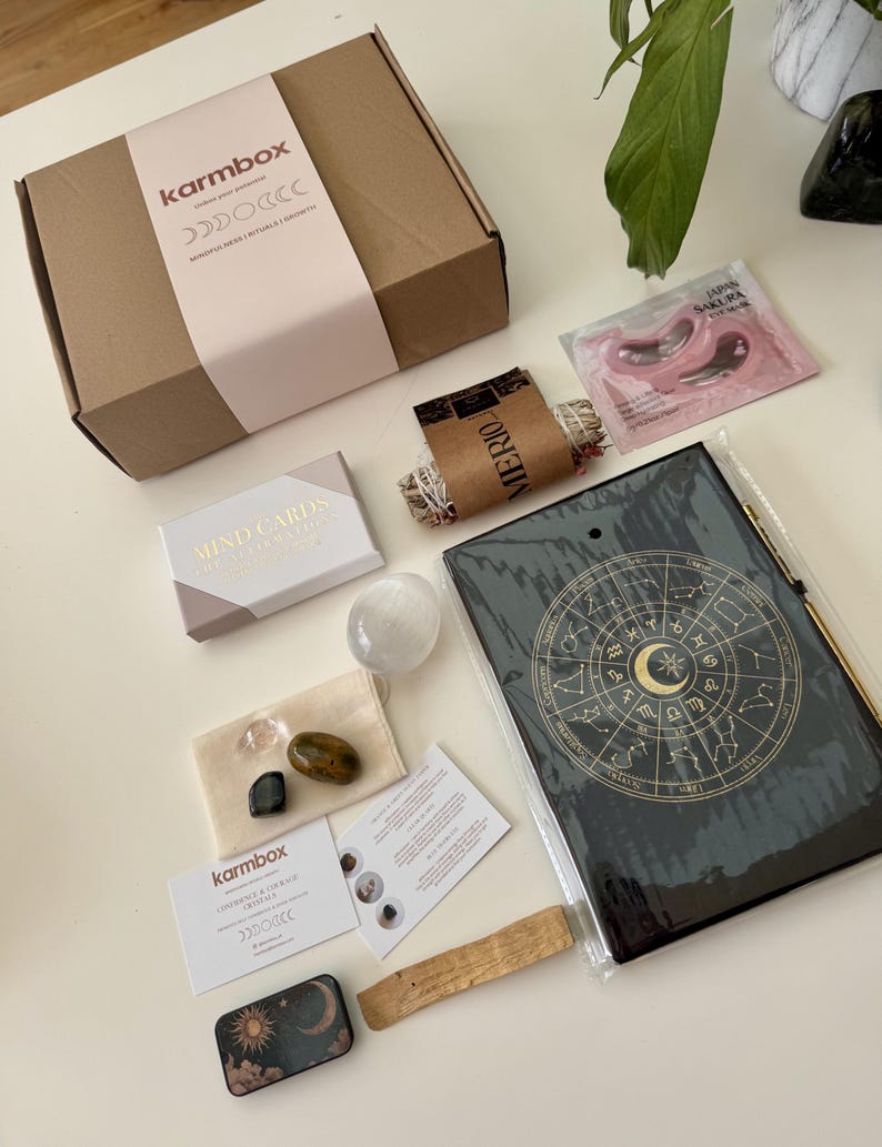 May include: A Karmbox cardboard shipping box, a pack of Japan Sakura eye masks, a sage bundle, a Mind Cards deck, a selenite stone, a small pouch with three crystals, a Karmbox card, a black zodiac journal, a palo santo stick, and a small sun and moon tin.