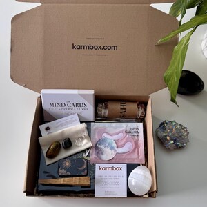 May include: A cardboard subscription box labeled karmbox.com containing LSW Mind Cards, a sage bundle, a small pouch with three polished stones, Japan Sakura eye masks, a moon-themed tin, a wooden stick, a white egg-shaped stone, and a card.