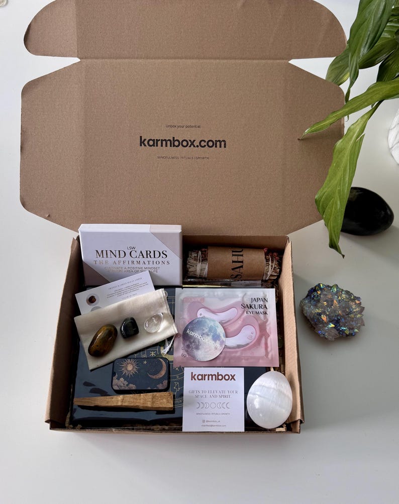 May include: A cardboard subscription box labeled karmbox.com containing LSW Mind Cards, a sage bundle, a small pouch with three polished stones, Japan Sakura eye masks, a moon-themed tin, a wooden stick, a white egg-shaped stone, and a card.