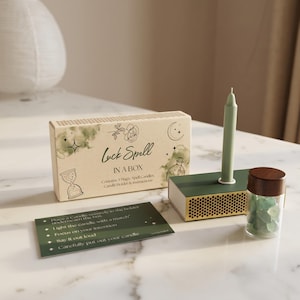May include: A Luck Spell in a Box set, featuring a box with text and illustrations, a green candle, a candle holder, a glass jar with a wooden lid, and a card with instructions. The box reads "Luck Spell IN A BOX".