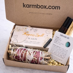 Może przedstawiać: Otwarty karton z napisem "karmbox.com". W środku zestaw "Happiness Spell in a Box", wiązka szałwii, bransoletka, mała butelka i karta "Labradorite".
