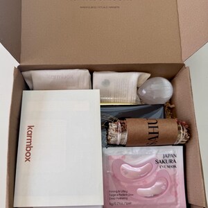 May include: A brown cardboard subscription box labeled karmbox.com containing a white information card, a Japan Sakura eye mask packet, a bundle of dried herbs wrapped in brown paper, a white crystal, and two small beige fabric pouches.