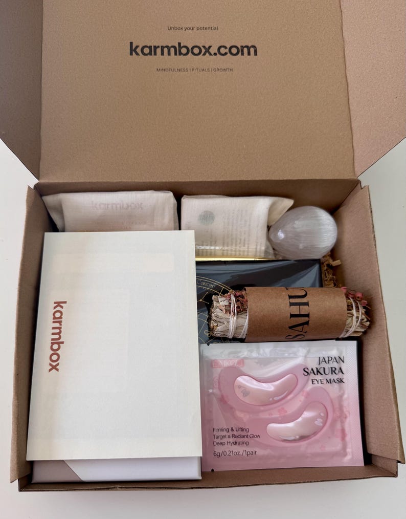 May include: A brown cardboard subscription box labeled karmbox.com containing a white information card, a Japan Sakura eye mask packet, a bundle of dried herbs wrapped in brown paper, a white crystal, and two small beige fabric pouches.