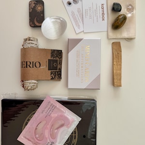 May include: A wellness kit containing a small metal tin with celestial art, a clear crystal sphere, a sage bundle, a deck of Mind Cards, a beige pouch with three stones, a palo santo stick, and a black book with pink Sakura eye masks.