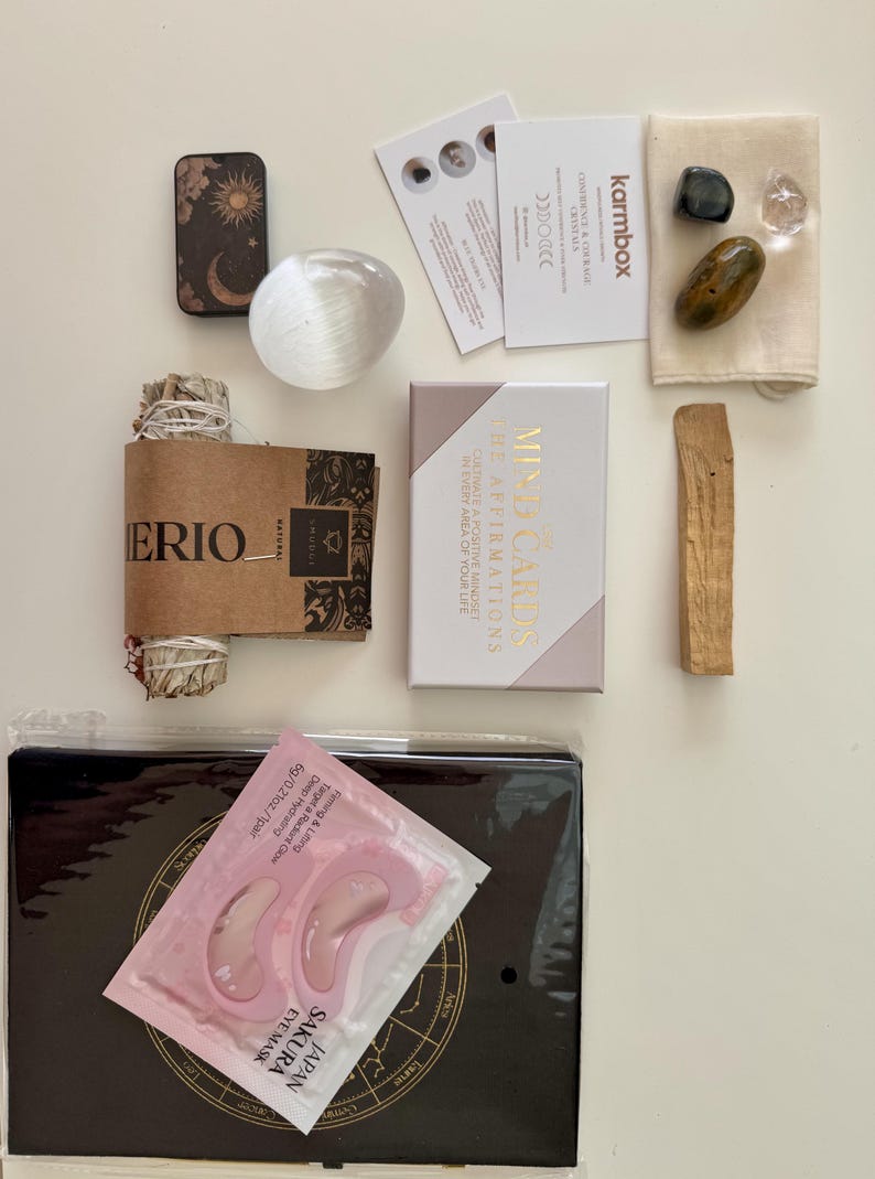 May include: A wellness kit containing a small metal tin with celestial art, a clear crystal sphere, a sage bundle, a deck of Mind Cards, a beige pouch with three stones, a palo santo stick, and a black book with pink Sakura eye masks.