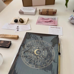 May include: A black notebook with a gold zodiac wheel design, a gold pen, a cardboard box labeled karmbox, a small box of mind cards, a bundle of dried herbs, a pink eye mask packet, two small stones in a pouch, and two informational cards.